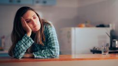A woman wearing pajamas leaning on her kitchen counter with her eyes closed